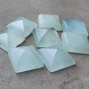 May include: A cluster of pale blue, pyramid-shaped gemstones. Each gem has a square base and a pointed top, with facets that reflect light. The stones are arranged on a textured, grey surface.