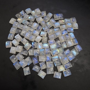 May include: A collection of square-cut moonstone gemstones. The stones are mostly white with iridescent blue and yellow flashes. The gemstones are scattered on a dark surface, showcasing their polished surfaces and natural variations.