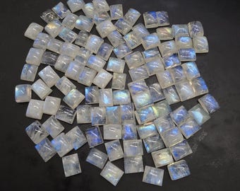 May include: A collection of square-cut moonstone gemstones. The stones are mostly white with iridescent blue and yellow flashes. The gemstones are scattered on a dark surface, showcasing their polished surfaces and natural variations.