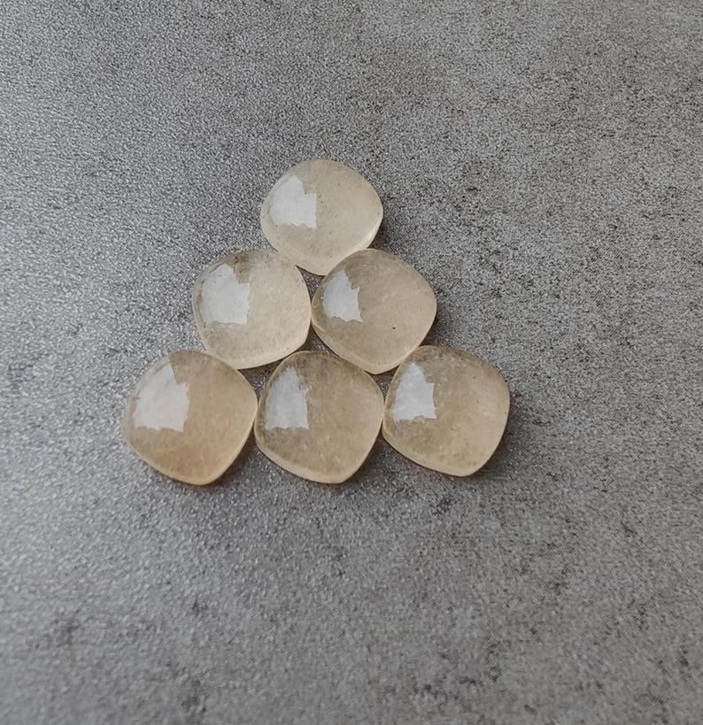 May include: Six light amber-colored, square-shaped gemstones arranged in a triangular pattern. The stones have rounded corners and a translucent quality, with a subtle sheen. They are displayed on a gray, textured surface.