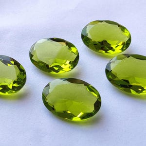 Op de afbeelding: Vijf ovale, groene edelstenen met een facetgeslepen vorm. De edelstenen zijn in een halve cirkel op een wit oppervlak gerangschikt.