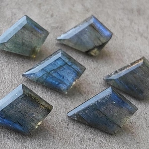 May include: Six trapezoid-cut labradorite gemstones in shades of blue and grey. The stones exhibit a shimmering play of colour. They are displayed on a neutral surface, highlighting their natural beauty and unique facets.