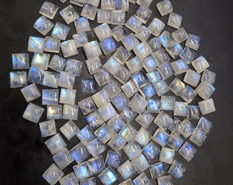 Könnte beinhalten: Eine Ansammlung quadratisch geschliffener Mondstein-Edelsteine. Die Steine sind überwiegend weiß mit irisierenden blauen und silbernen Reflexen. Die Edelsteine sind auf einer dunklen Oberfläche verstreut, was ihre natürliche Schönheit hervorhebt.