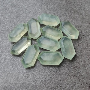 May include: A collection of pale green, faceted gemstone crystals with a hexagonal shape. The crystals are arranged on a gray surface, showcasing their translucent quality and geometric design. The gemstones have a polished appearance.