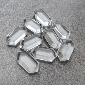 May include: A cluster of clear, faceted crystal gemstones. Each gemstone has a rectangular shape with beveled edges. They are scattered on a grey surface, reflecting light. Suitable for jewellery making or crafts.