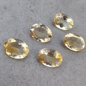 Può includere: Cinque pietre preziose ovali, sfaccettate, di colore giallo chiaro. Le pietre preziose sono disposte a semicerchio su una superficie grigia.