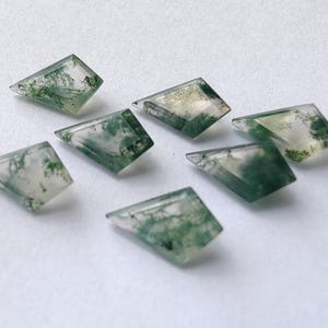 May include: Seven translucent, geometric-shaped moss agate gemstones. Each stone features a unique pattern of green, fern-like inclusions within a clear, crystalline structure. The gemstones are arranged on a white surface.
