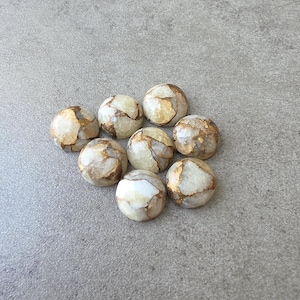 May include: A collection of eight round, polished stones with a marbled appearance. The stones have a creamy white base color with veins of gold and brown. The stones are arranged on a gray surface.