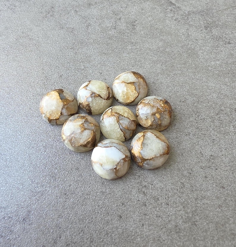 May include: A collection of eight round, polished stones with a marbled appearance. The stones have a creamy white base color with veins of gold and brown. The stones are arranged on a gray surface.
