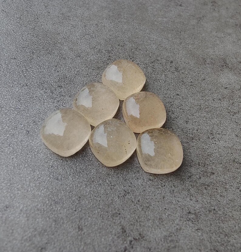 May include: Six light amber-colored, square-shaped gemstones arranged in a triangular pattern. The stones have rounded corners and a translucent quality, with a subtle sheen. The background is a mottled gray surface.