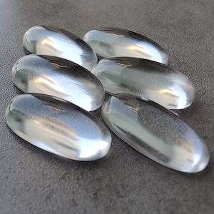 May include: Six clear, oval-shaped, smooth glass stones. The stones are arranged in a cluster on a grey surface.