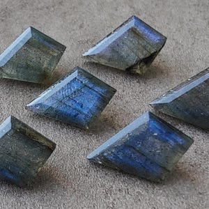 May include: Six faceted labradorite gemstones in shades of blue, green, and grey. The gemstones are cut into rectangular shapes with angled edges, showing a polished, reflective surface. The stones are arranged on a textured, neutral-toned surface.