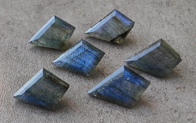 May include: Six faceted labradorite gemstones in shades of blue, green, and grey. The gemstones are cut into rectangular shapes with angled edges, showing a polished, reflective surface. The stones are arranged on a textured, neutral-toned surface.