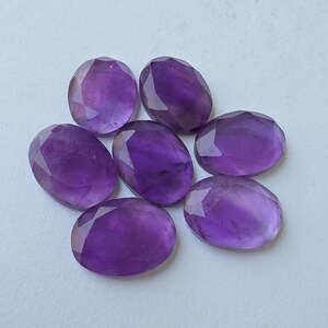 May include: Seven faceted oval amethyst gemstones in varying shades of purple. The gemstones are arranged in a circular pattern, each with a polished surface and a brilliant cut. The stones are displayed on a white surface.