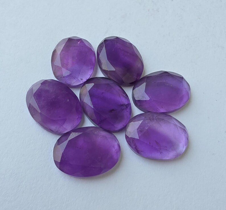 May include: Seven faceted oval amethyst gemstones in varying shades of purple. The gemstones are arranged in a circular pattern, each with a polished surface and a brilliant cut. The stones are displayed on a white surface.