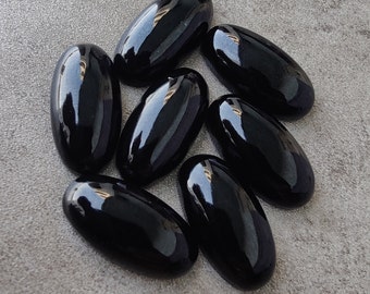 May include: Seven black oval-shaped gemstones, likely obsidian, arranged in a cluster on a gray surface.