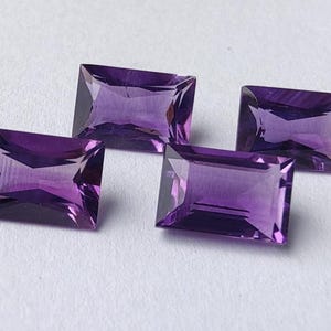 Può includere: Quattro pietre preziose di ametista rettangolari e sfaccettate sono esposte su uno sfondo bianco. Le gemme hanno una tonalità viola intenso e sono tagliate con angoli precisi, riflettendo la luce. Le pietre preziose sono disposte in modo leggermente sparso.