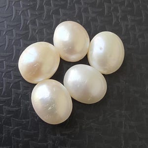 Op de afbeelding: Vijf ovale parels in een cr&egrave;mewitte kleur liggen op een donkere, getextureerde ondergrond. De parels hebben een glad, reflecterend oppervlak, dat het licht opvangt en subtiele iriserende effecten vertoont. Deze edelstenen zijn ideaal voor het maken van sieraden.