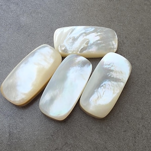 Puede incluir: Cuatro cabujones rectangulares de nácar con bordes redondeados. Las piedras iridiscentes blancas y crema están dispuestas sobre una superficie gris. Los cabujones tienen una superficie lisa y pulida que refleja la luz.