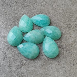 Op de afbeelding: Zeven gefacetteerde, druppelvormige edelstenen in tinten turquoise en zeegroen liggen op een getextureerd grijs oppervlak. De edelstenen hebben een gepolijste, glanzende uitstraling met meerdere facetten.
