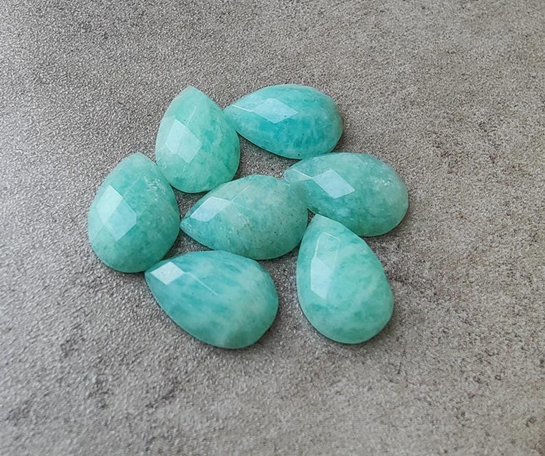Op de afbeelding: Zeven gefacetteerde, druppelvormige edelstenen in tinten turquoise en zeegroen liggen op een getextureerd grijs oppervlak. De edelstenen hebben een gepolijste, glanzende uitstraling met meerdere facetten.