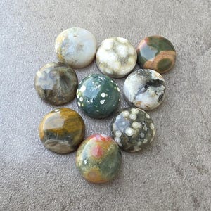 May include: A collection of nine polished, round gemstones in various colors and patterns. The stones include shades of green, brown, white, and red, with unique veining and speckled designs. The gemstones are arranged on a gray surface.