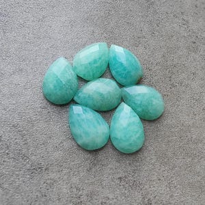 Op de afbeelding: Zeven gefacetteerde, druppelvormige Amazoniet edelstenen in verschillende maten. De stenen zijn licht turquoise van kleur met subtiele adering en een gepolijst, reflecterend oppervlak. Ze zijn gerangschikt op een getextureerd, grijs oppervlak.