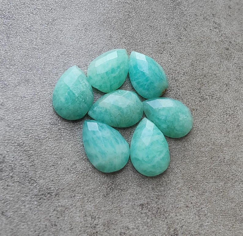 Op de afbeelding: Zeven gefacetteerde, druppelvormige Amazoniet edelstenen in verschillende maten. De stenen zijn licht turquoise van kleur met subtiele adering en een gepolijst, reflecterend oppervlak. Ze zijn gerangschikt op een getextureerd, grijs oppervlak.