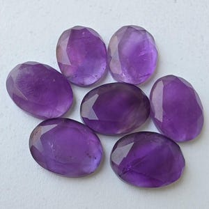 May include: Seven faceted, oval-shaped amethyst gemstones in varying shades of purple are arranged on a white surface. The gemstones have a polished, reflective surface, showcasing their natural color and clarity. The arrangement is symmetrical.