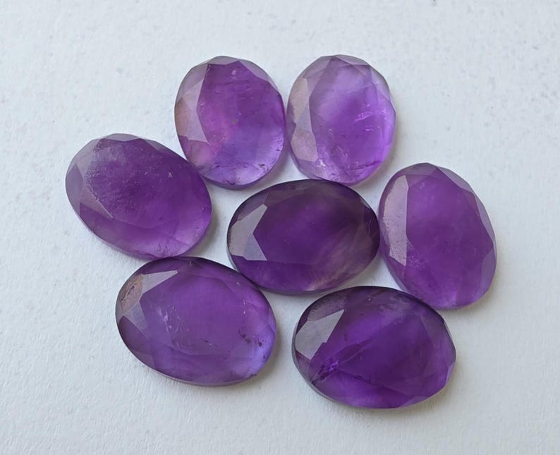 May include: Seven faceted, oval-shaped amethyst gemstones in varying shades of purple are arranged on a white surface. The gemstones have a polished, reflective surface, showcasing their natural color and clarity. The arrangement is symmetrical.