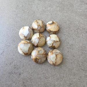 May include: Nine round, polished stones with a cream and gold color scheme. The stones have a marbled appearance with gold veining throughout. The stones are arranged in a square pattern on a gray surface.