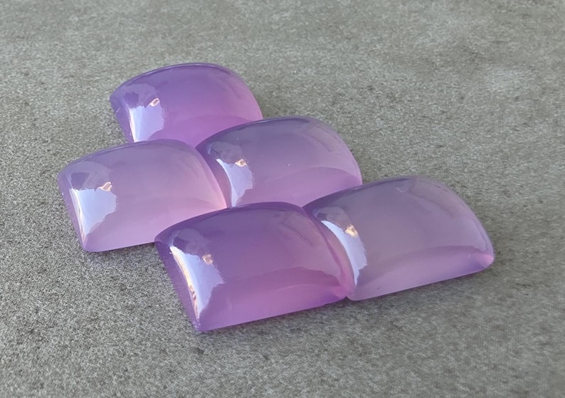 Pu&ograve; includere: Cinque gemme lucidate, a taglio quadrato, in varie tonalit&agrave; di lavanda e viola. Le pietre sono disposte in modo leggermente sovrapposto, con una superficie lucida e riflettente. Le gemme sono su uno sfondo grigio neutro.