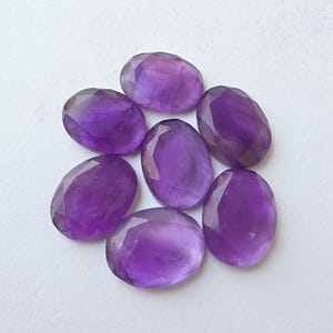 May include: Seven faceted oval amethyst gemstones in varying shades of purple. The gemstones are arranged in a circular pattern, showcasing their polished surfaces and internal clarity. Ideal for jewelry making or collecting.