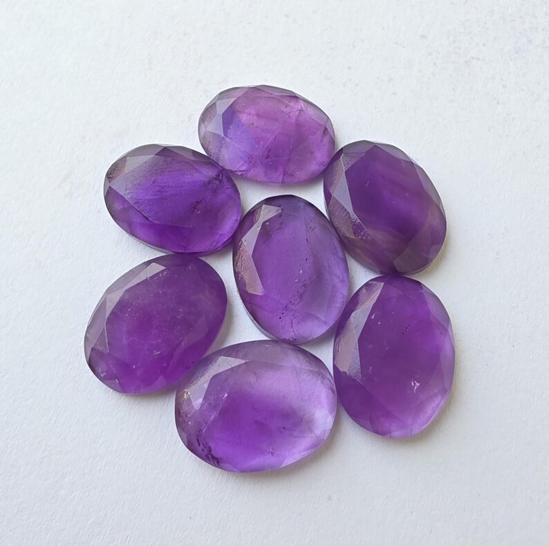 May include: Seven faceted oval amethyst gemstones in varying shades of purple. The gemstones are arranged in a circular pattern, showcasing their polished surfaces and internal clarity. Ideal for jewelry making or collecting.