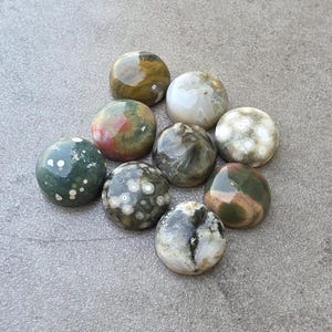 May include: A collection of nine polished, spherical gemstones. The stones display a variety of colors, including green, brown, white, and red, with unique patterns and textures. The gemstones are arranged on a gray surface.