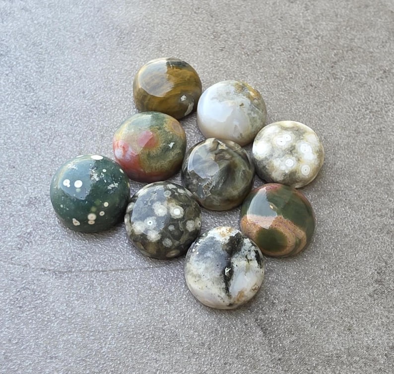 May include: A collection of nine polished, spherical gemstones. The stones display a variety of colors, including green, brown, white, and red, with unique patterns and textures. The gemstones are arranged on a gray surface.