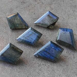 May include: Six faceted labradorite gemstones in shades of blue and grey. Each stone has a distinct shape and polished surface, reflecting light. The gemstones are arranged on a grey surface, highlighting their natural beauty.
