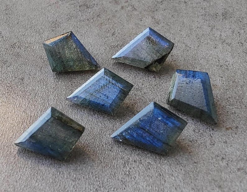 May include: Six faceted labradorite gemstones in shades of blue and grey. Each stone has a distinct shape and polished surface, reflecting light. The gemstones are arranged on a grey surface, highlighting their natural beauty.