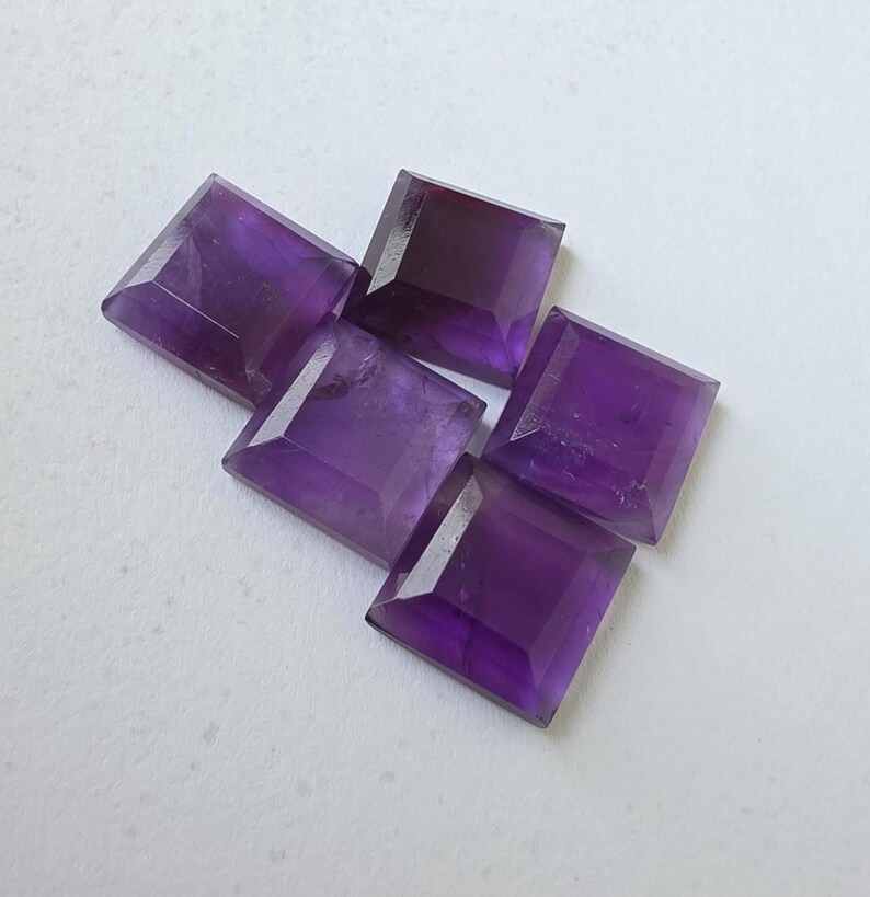 Peut inclure: Cinq pierres pr&eacute;cieuses d'am&eacute;thyste taill&eacute;es en carr&eacute;, de diff&eacute;rentes nuances de violet, sont dispos&eacute;es sur une surface blanche. Les pierres ont un design &agrave; facettes, captant la lumi&egrave;re et mettant en valeur leur couleur riche. Ces pierres sont id&eacute;ales pour la joaillerie.