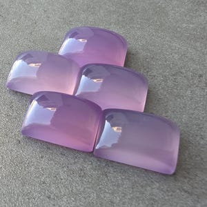 Pu&ograve; includere: Cinque pietre preziose lucide color lavanda con bordi arrotondati sono disposte su una superficie grigia. Le pietre hanno una qualit&agrave; traslucida, riflettendo la luce. Questi cabochon sono ideali per la creazione di gioielli. Ogni pietra misura circa 2 cm.