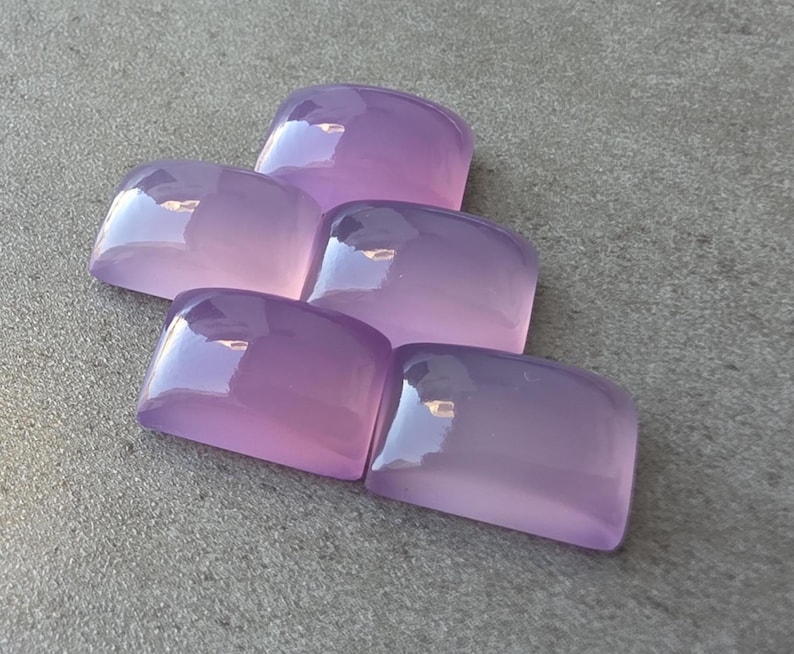 Pu&ograve; includere: Cinque pietre preziose lucide color lavanda con bordi arrotondati sono disposte su una superficie grigia. Le pietre hanno una qualit&agrave; traslucida, riflettendo la luce. Questi cabochon sono ideali per la creazione di gioielli. Ogni pietra misura circa 2 cm.