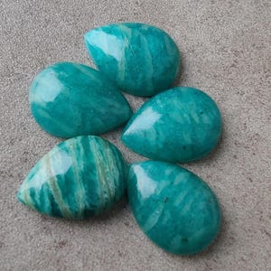 Pode incluir: Cinco pedras preciosas de amazonita polidas em tons de verde-azulado e turquesa. As pedras têm formato de gota, com uma superfície lisa e brilhante. Algumas pedras apresentam veios visíveis em verde claro e branco.