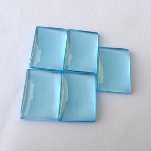 May include: Five pale blue glass tiles arranged together. Each tile is square with one angled edge. The tiles are translucent, reflecting light. These tiles are likely for mosaic or craft projects. Each tile is approximately 2.5 cm square.