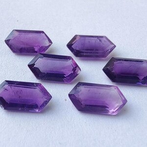 K&ouml;nnte beinhalten: Sechs facettierte, sechseckige Amethyst-Edelsteine in verschiedenen Lilat&ouml;nen liegen auf einer wei&szlig;en Oberfl&auml;che. Die Edelsteine haben eine polierte, reflektierende Oberfl&auml;che und sind mit flachen Ober- und Unterseiten geschnitten.