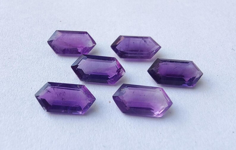 K&ouml;nnte beinhalten: Sechs facettierte, sechseckige Amethyst-Edelsteine in verschiedenen Lilat&ouml;nen liegen auf einer wei&szlig;en Oberfl&auml;che. Die Edelsteine haben eine polierte, reflektierende Oberfl&auml;che und sind mit flachen Ober- und Unterseiten geschnitten.