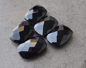 May include: Five faceted, black gemstone beads are arranged on a grey surface. The beads have a geometric shape with multiple flat surfaces that reflect light. The gemstones are opaque and have a polished finish.