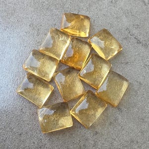 May include: A collection of eleven amber-colored, rectangular glass tiles. Each tile has rounded corners and a slightly translucent appearance, with variations in the intensity of the amber hue. The tiles are arranged on a gray surface.
