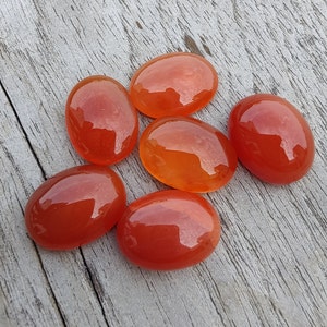 Puede incluir: Seis piedras de cornalina lisas de forma ovalada en tonos naranja y rojo. Las piedras están dispuestas sobre una superficie de madera marrón claro.