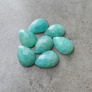 May include: Seven faceted, teardrop-shaped Amazonite gemstones in varying sizes are arranged on a grey surface. The stones display a light turquoise colour with subtle variations in tone. Each stone has a polished, reflective surface.
