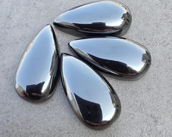 May include: Four polished, teardrop-shaped stones with a reflective, dark gray surface. The stones are arranged on a light gray surface, with each stone reflecting the surrounding environment. The smooth, glossy finish suggests a natural material.